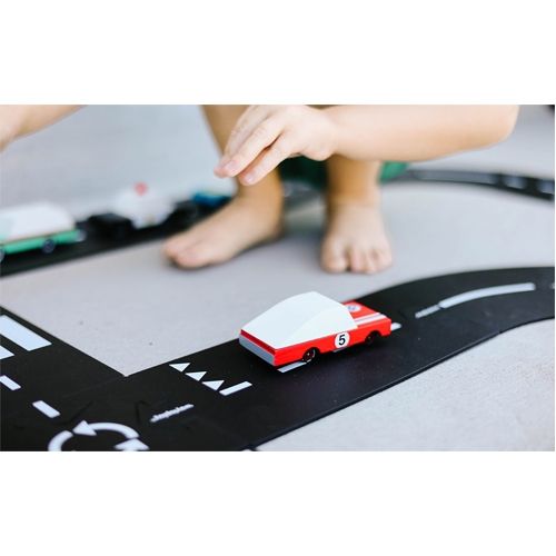 Een Candylab houten speelgoedauto rijdt op een zwarte speelmat met wegmarkeringen, met kindervoeten op de achtergrond.