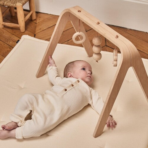 Een baby ligt op een speelmat en reikt naar de Charlie Crane houten babygym.