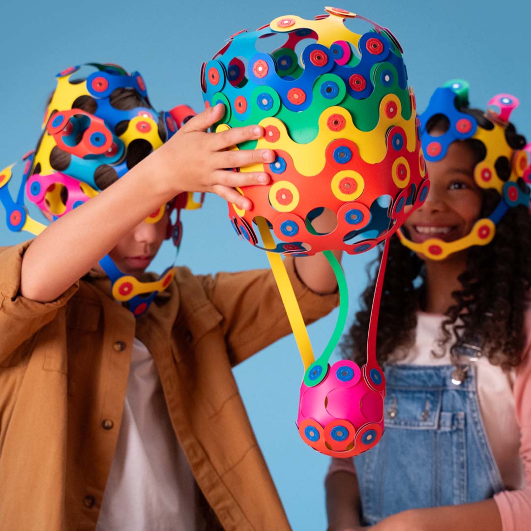 Twee kinderen spelen met kleurrijke Clixo bouwsels, waaronder een grote bol en een hoofddeksel.