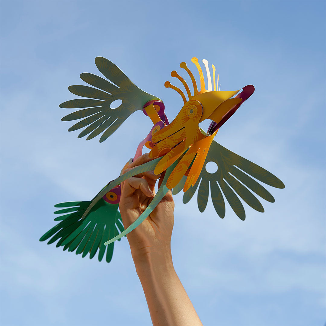 Een hand houdt een kleurrijke tropische vogel van de Clixo magnetische bouwset omhoog tegen een blauwe lucht.