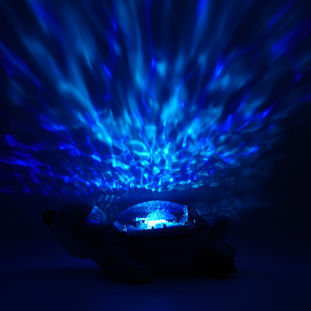 cloud.b knuffelpinguin met nachtlamp die blauwe watergolven projecteert op een plafond in het donker.