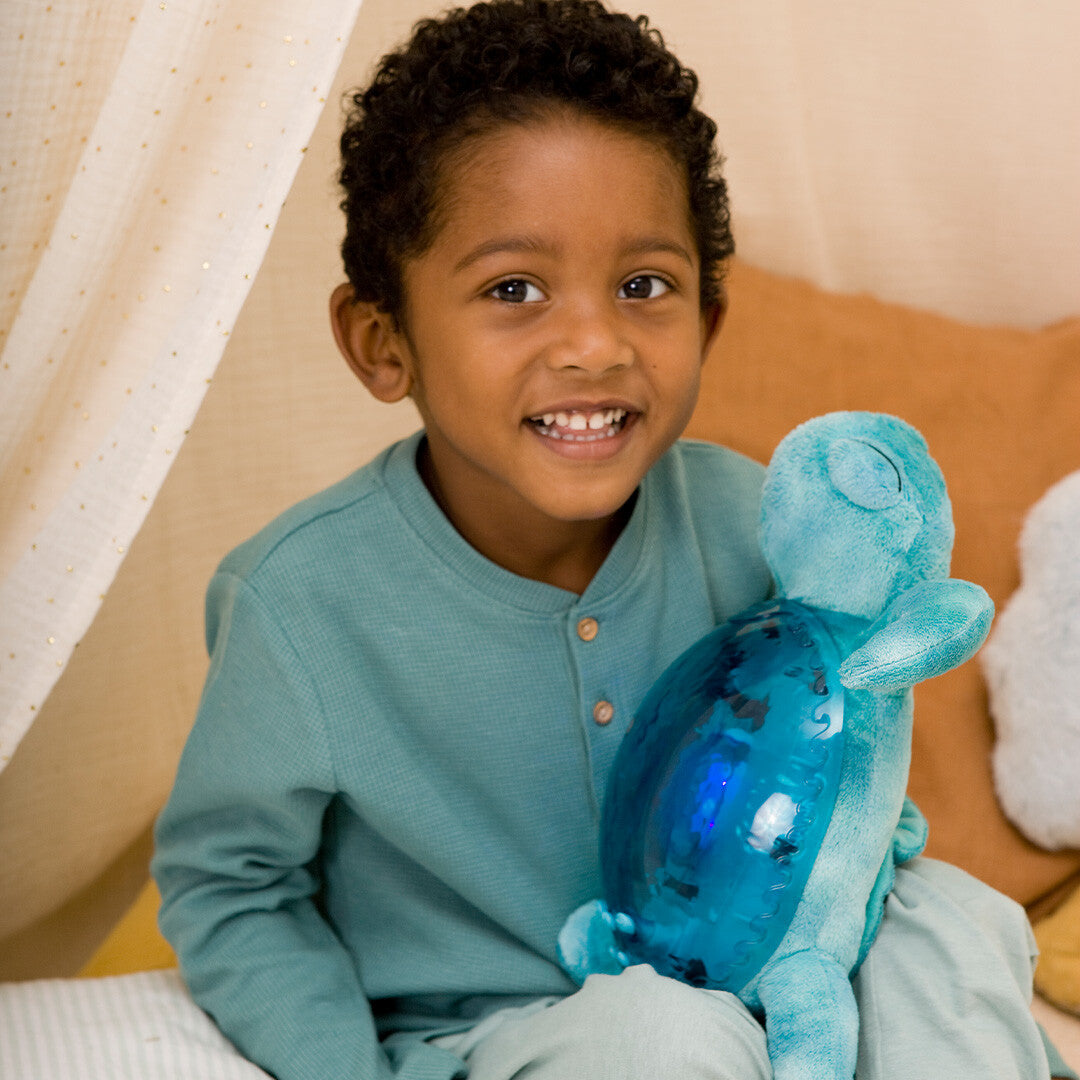 Een lachende jongen zit met de cloud.b knuffelschildpad met nachtlamp op schoot.