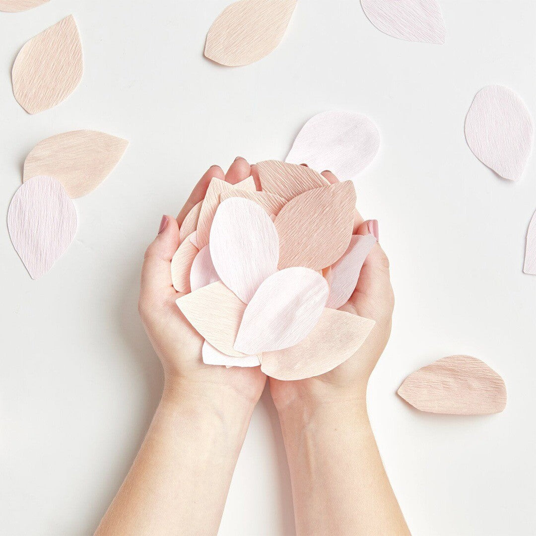 Twee handen houden Rico Design confetti rozenblaadjes vast, met enkele blaadjes verspreid op een witte ondergrond.