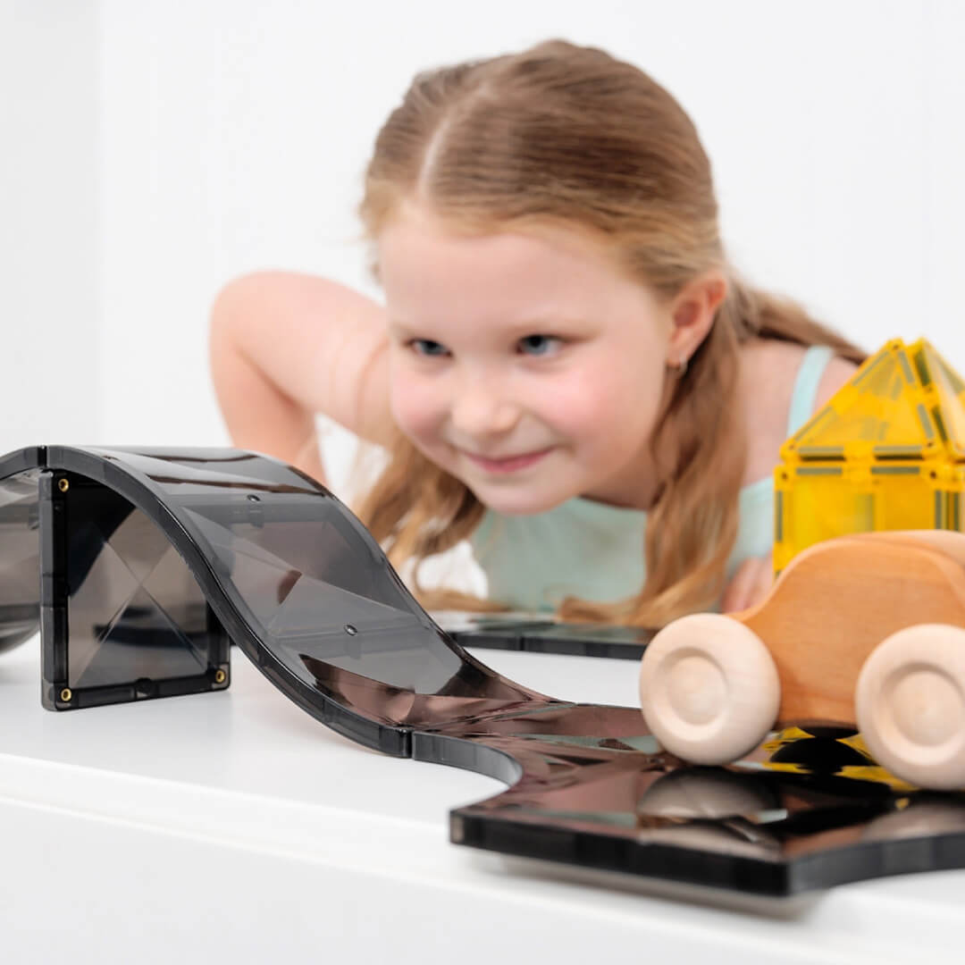 Een kind kijkt lachend naar een houten auto op een helling van zwarte Connetix magnetische tegels.