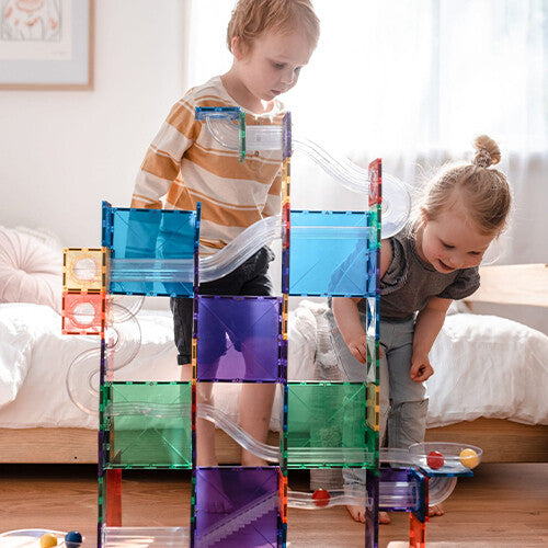 Twee jonge kinderen kijken aandachtig naar een Connetix knikkerbaan, gebouwd met kleurrijke magnetische tegels en transparante banen.