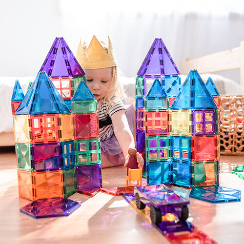 Een kind met een kroon speelt op de vloer met twee grote, kleurrijke Connetix magnetische tegelkastelen en een klein voertuig.