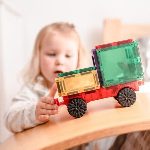 Een jong kind speelt met een rode en groene Connetix magnetische tegelvrachtwagen op een houten balansbord.