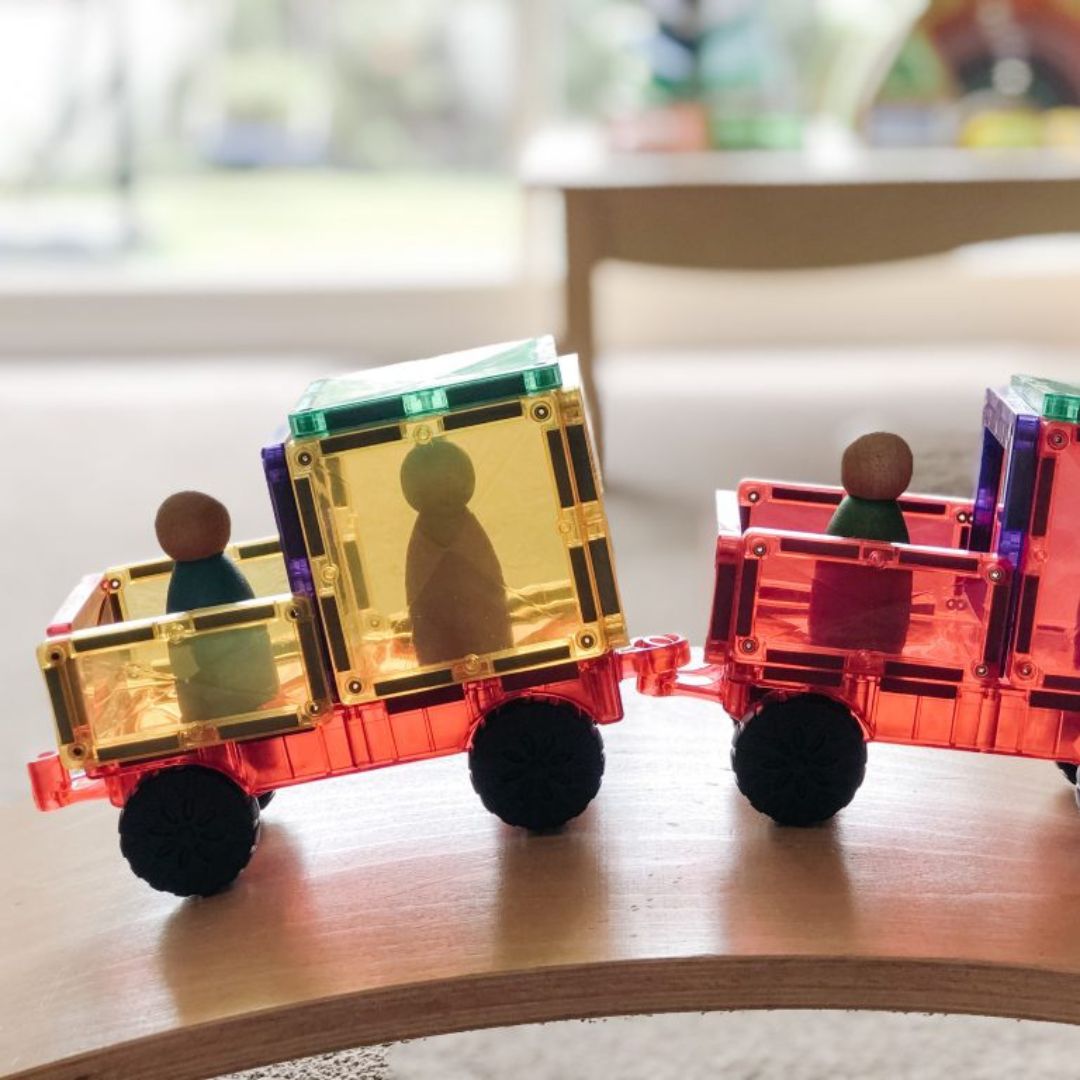 Een Connetix magnetische tegelvrachtwagen met aanhanger en houten poppetjes, rijdend op een houten balansbord.