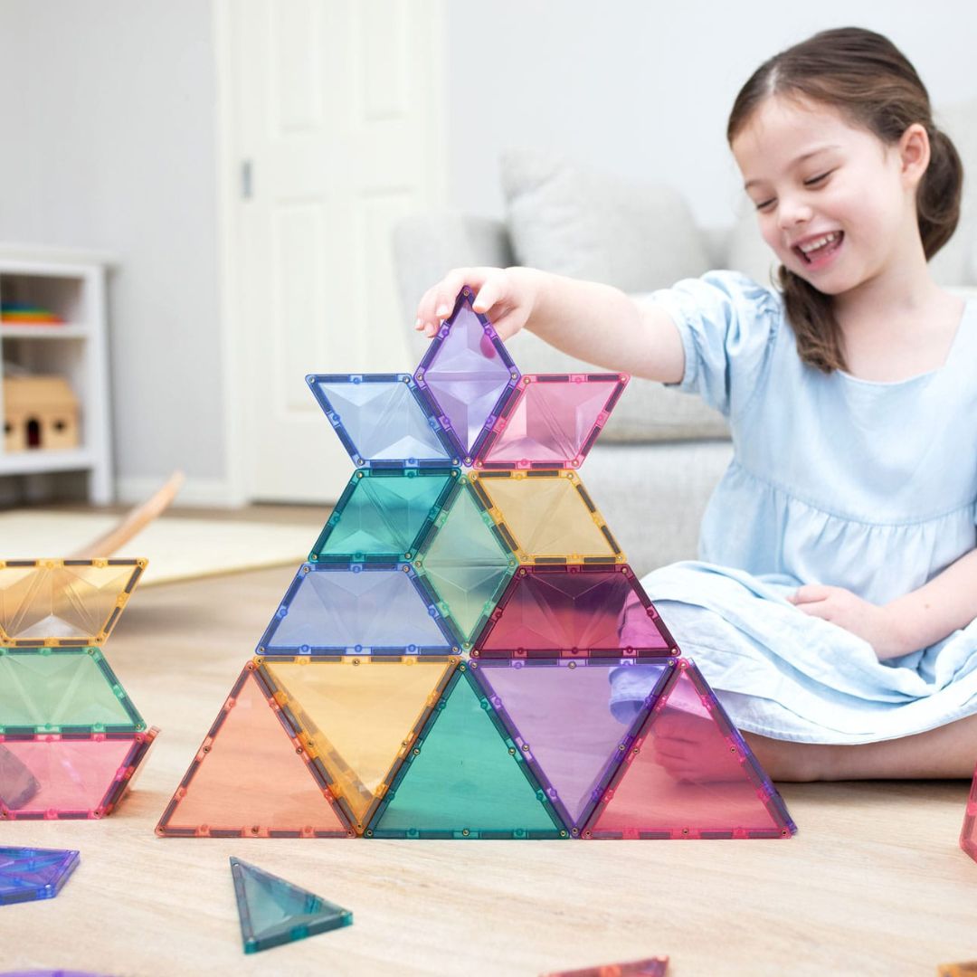 Een lachend meisje bouwt een piramide van Connetix pastel magnetische tegels op de vloer.