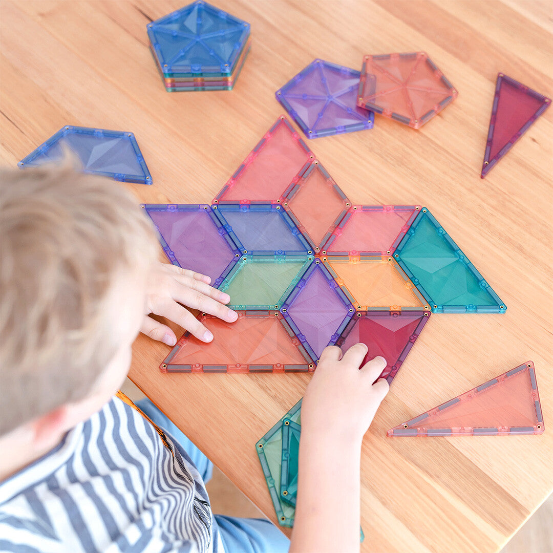 Een kind legt Connetix pastel magnetische tegels in een patroon op een houten tafel.
