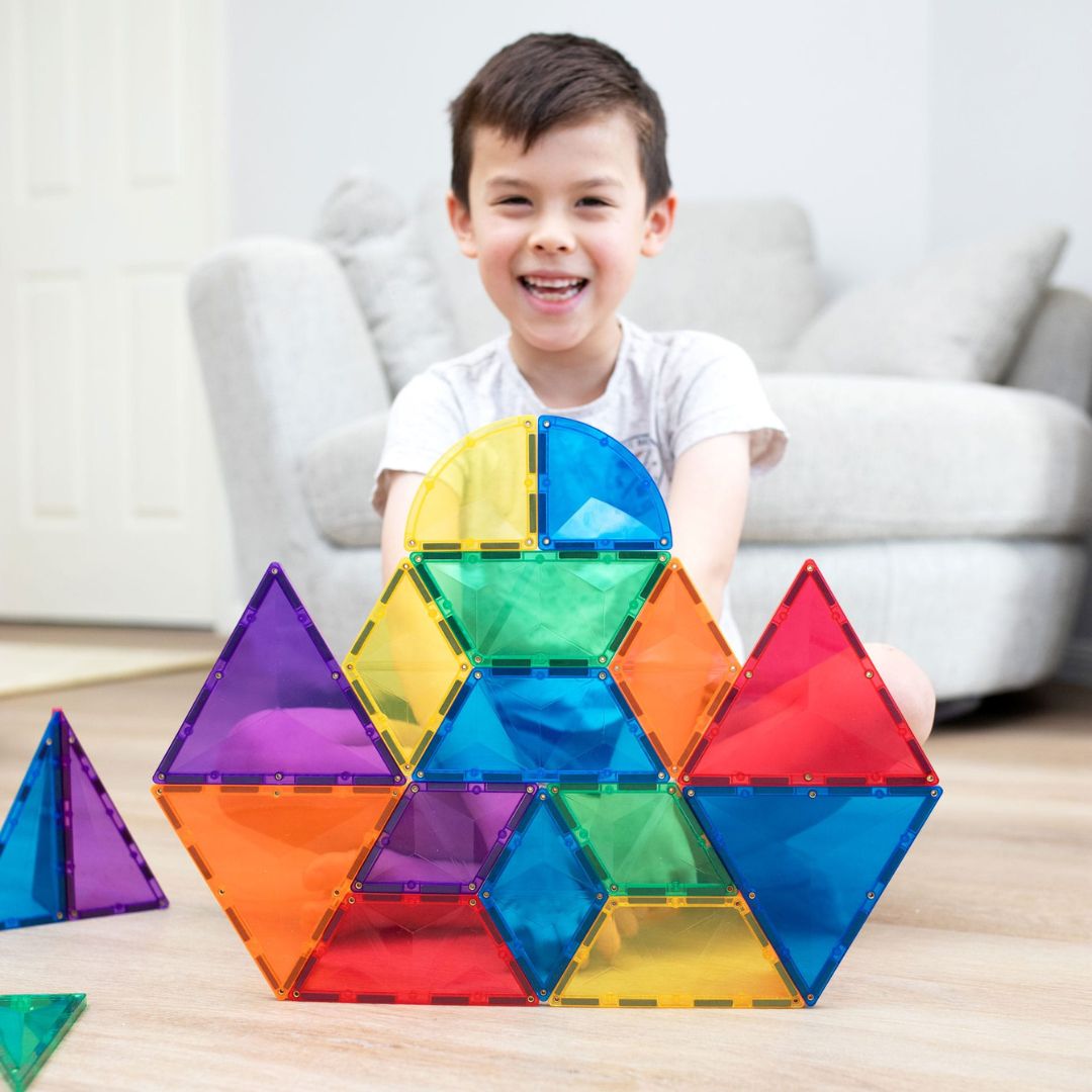 Een lachend kind zit achter een grote constructie van Connetix magnetische tegels in regenboogkleuren.
