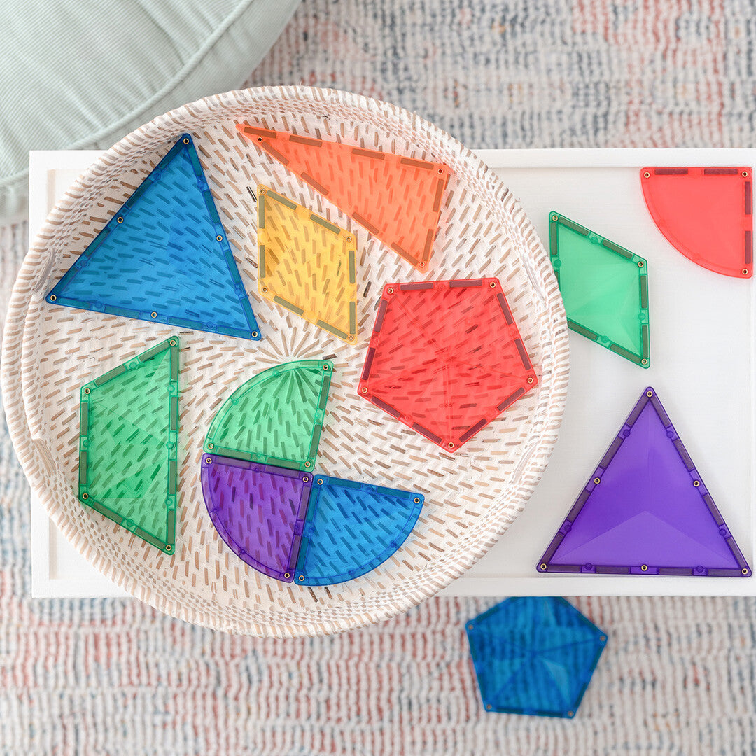 Een collectie Connetix magnetische tegels in diverse vormen en regenboogkleuren ligt in een mand en op een tafel.