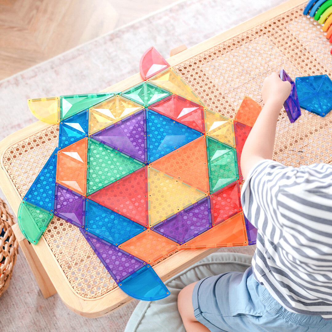 Een kind bouwt een grote platte figuur van Connetix magnetische tegels in regenboogkleuren op een rieten tafel.
