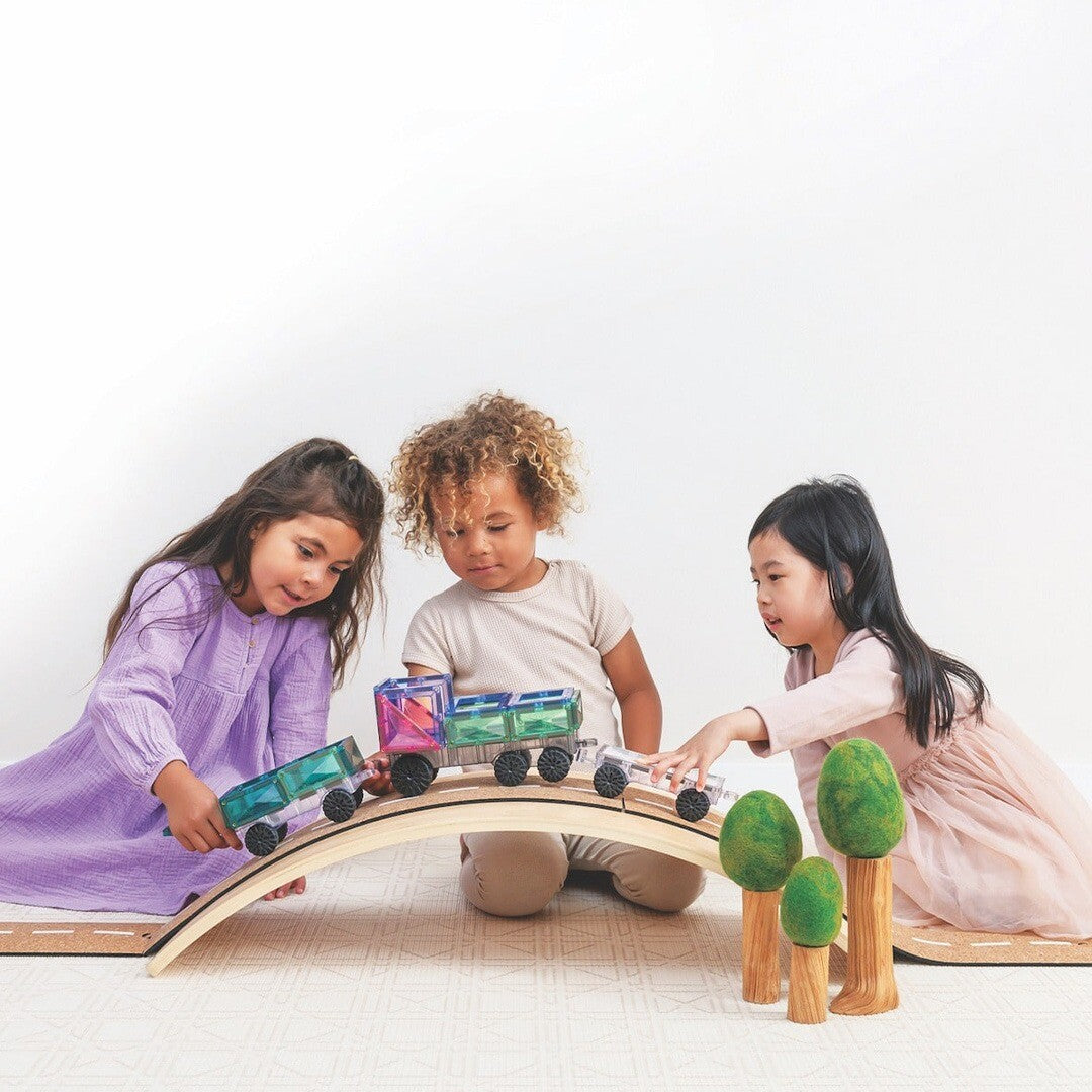 Drie kinderen spelen samen met een Connetix trein, gemaakt van pastelkleurige magnetische tegels, op een houten balansbord.