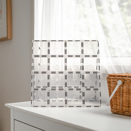 Een transparante Connetix magnetische bouwplaat staat rechtop op een witte kast voor een raam.