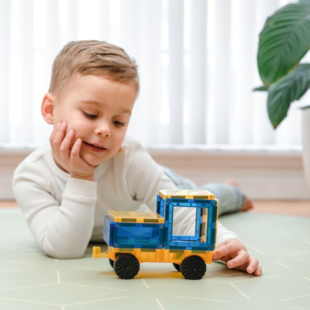 Een jongetje ligt op de grond en kijkt naar een Connetix auto, gemaakt van blauwe en gele magnetische tegels.