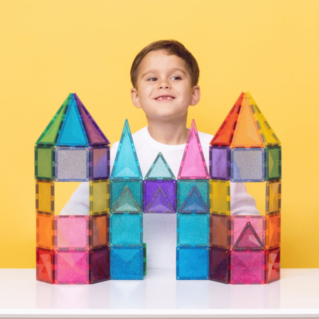 Een lachende jongen achter een kleurrijk kasteel gebouwd met Connetix glitter magnetische tegels.