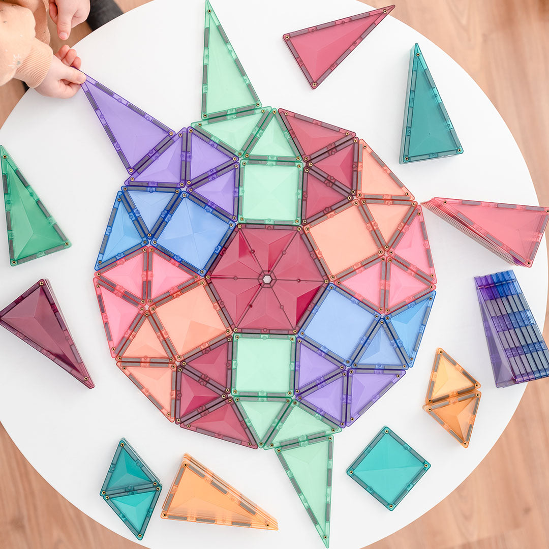 Een kind bouwt een groot geometrisch patroon met Connetix pastel magnetische tegels op een witte tafel.