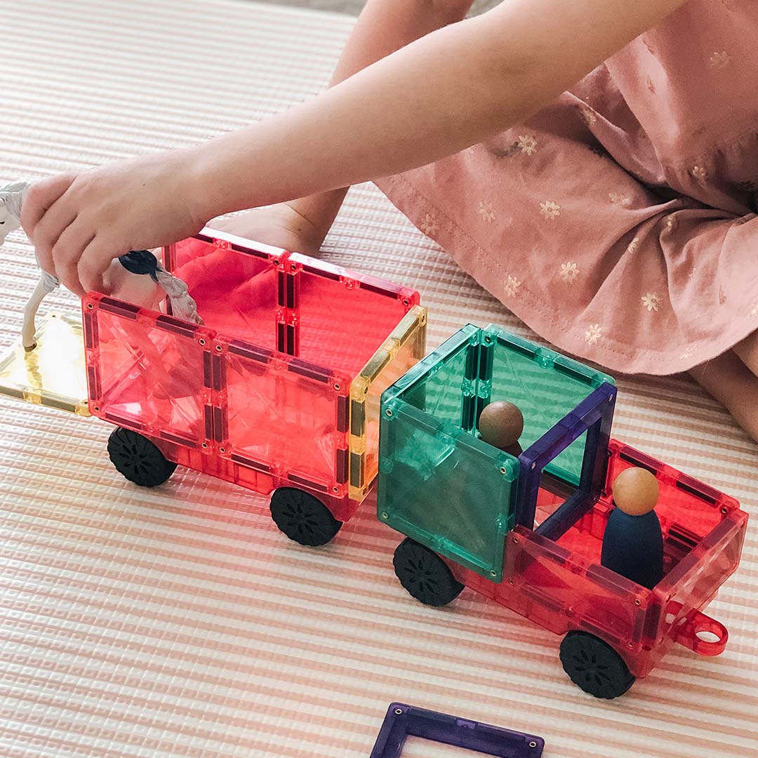 Kinderhanden spelen met een rode Connetix magnetische tegelvrachtwagen en aanhanger, met een speelgoedpaard in de aanhanger.