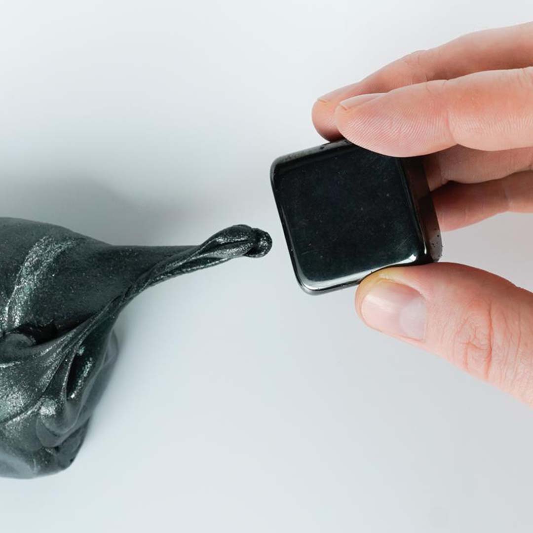 Een hand houdt een zwarte magneet dichtbij donkergrijze magnetische klei van Crazy Aaron's, die zich uitstrekt naar de magneet.