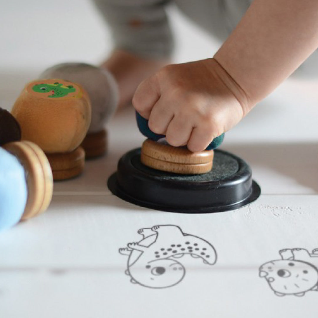Een kinderhand drukt een blauwe Crealign dinosaurusstempel in een zwart inktkussen, met gestempelde dino's op papier.
