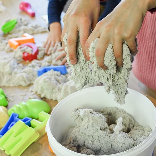 Kinderhanden spelen met Creall modelleerzand, dat uit elkaar getrokken wordt boven een witte emmer.