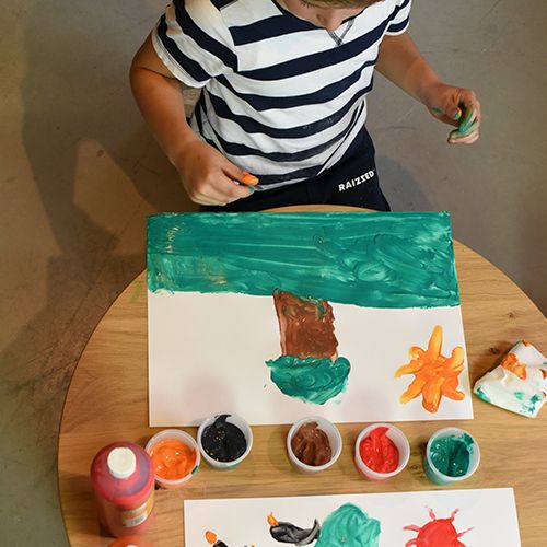 Een kind schildert met Creall vingerverf op een wit vel papier, met verschillende kleuren verf in potjes.