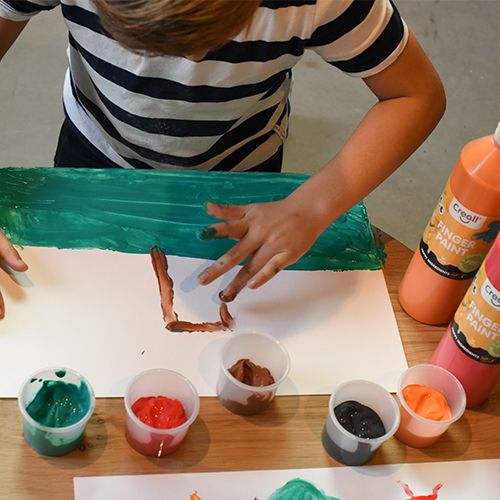Een kind schildert met Creall vingerverf op een vel papier, met flessen vingerverf op de achtergrond.