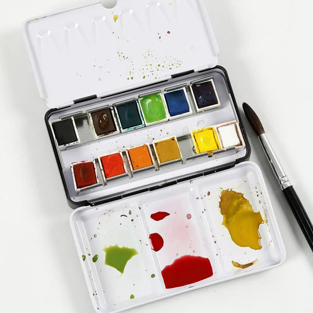 Een open metalen doos met 12 kleuren aquarelverf en een creativ company penseel ernaast.