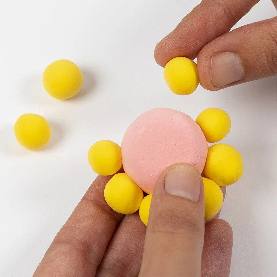 Handen vormen een bloem door gele balletjes Creativ Company Silk Clay aan een roze bal te bevestigen.