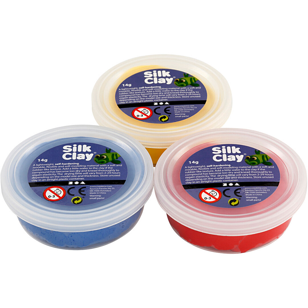 Drie potjes creativ company Silk Clay boetseerklei in geel, rood en blauw, schuin van bovenaf gefotografeerd.