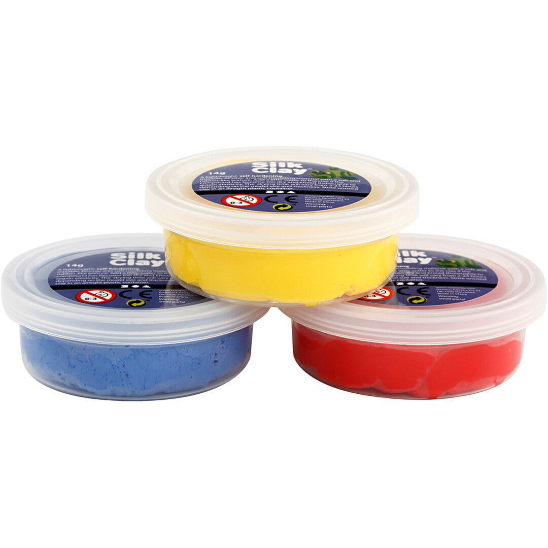 Drie potjes creativ company Silk Clay boetseerklei in geel, rood en blauw, schuin van voren gefotografeerd.