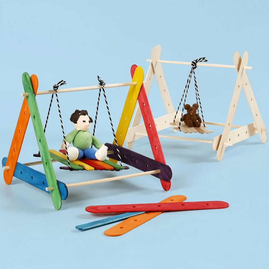 Twee zelfgemaakte schommels van houten constructiestokjes, één met een pop en één met een teddybeer, met losse gekleurde stokjes op de voorgrond.
