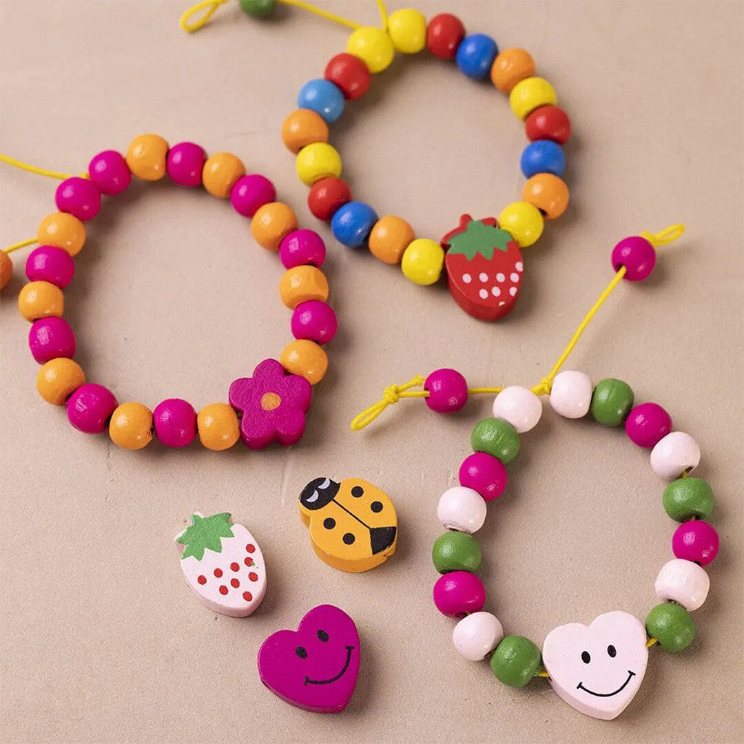 Drie kleurrijke kinderarmbandjes gemaakt van houten kralen met decoratieve bedels zoals een aardbei, bloem en hartje, naast losse kralen.