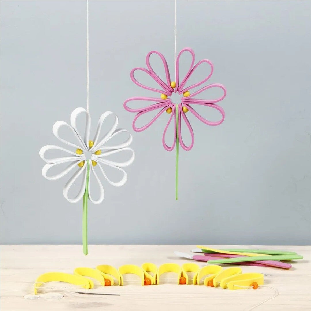 Twee hangende bloemen gemaakt van foam ijsstokjes, met extra gele foam stokjes en een naald op een houten tafel.