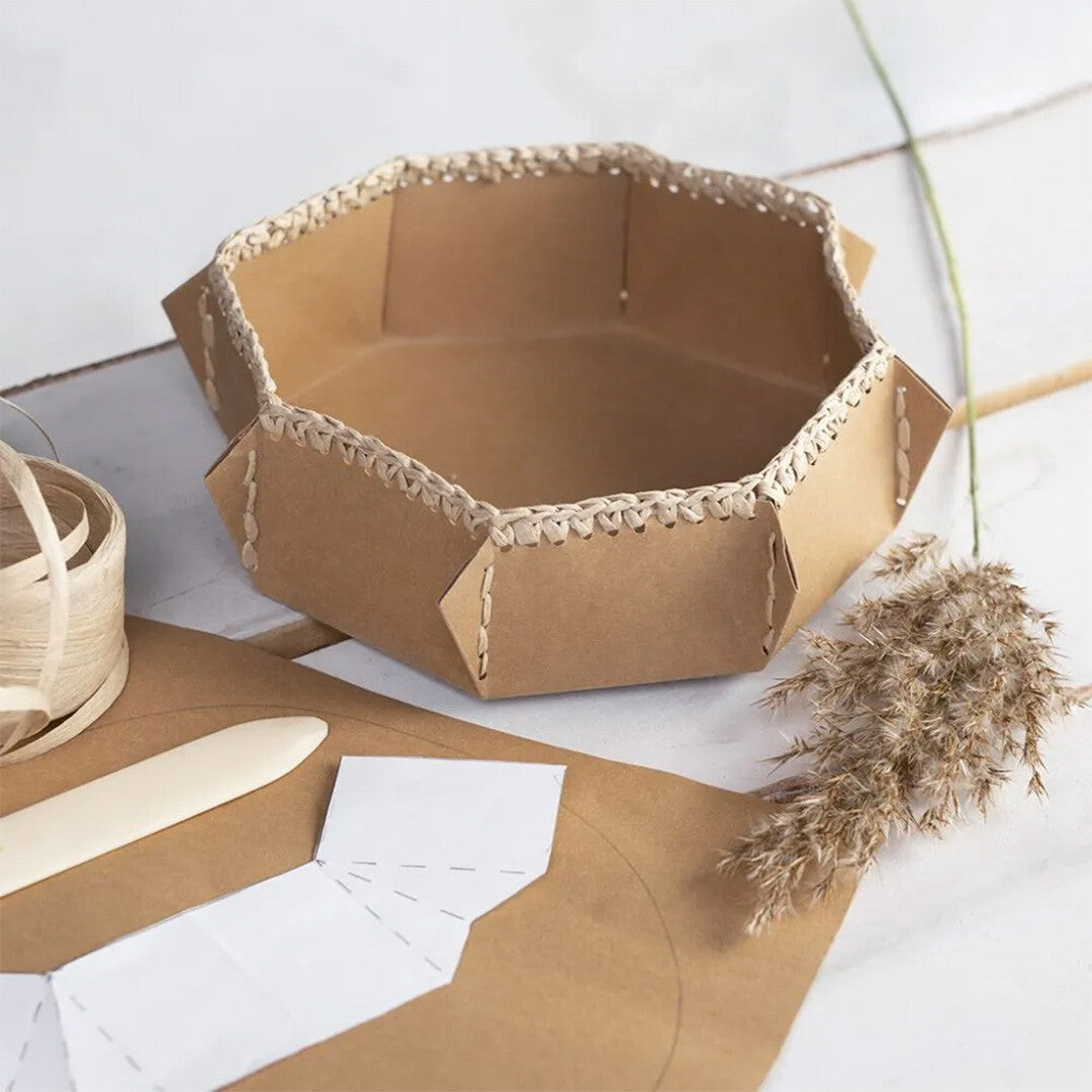 Een zelfgemaakte achthoekige mand van karton met raffia papiergaren details, naast knutselbenodigdheden.