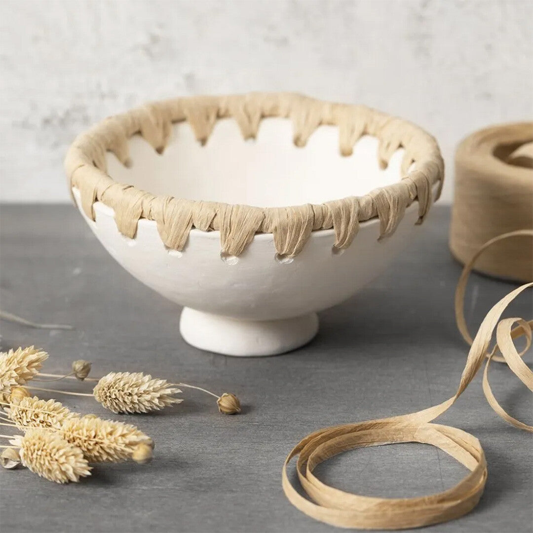 Een witte kom versierd met raffia papiergaren, naast gedroogde bloemen en losse raffia.