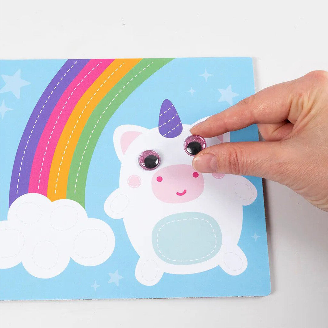 Een hand plakt een roze Creativ Company glitter wiebeloog op een tekening van een eenhoorn met een regenboog.