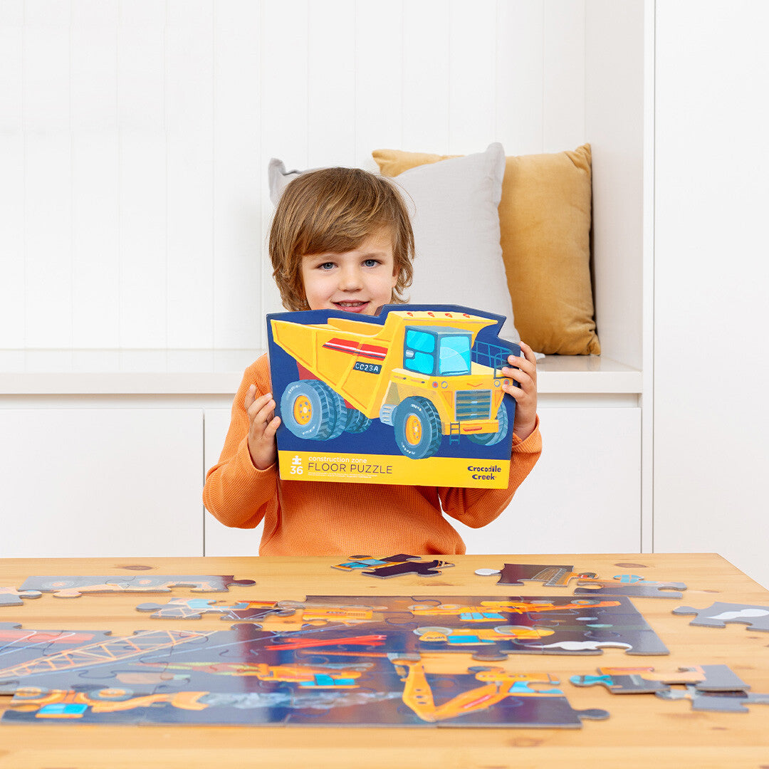 Een lachende jongen houdt de Crocodile Creek vloerpuzzel bouwplaats verpakking vast, met de puzzel op tafel.