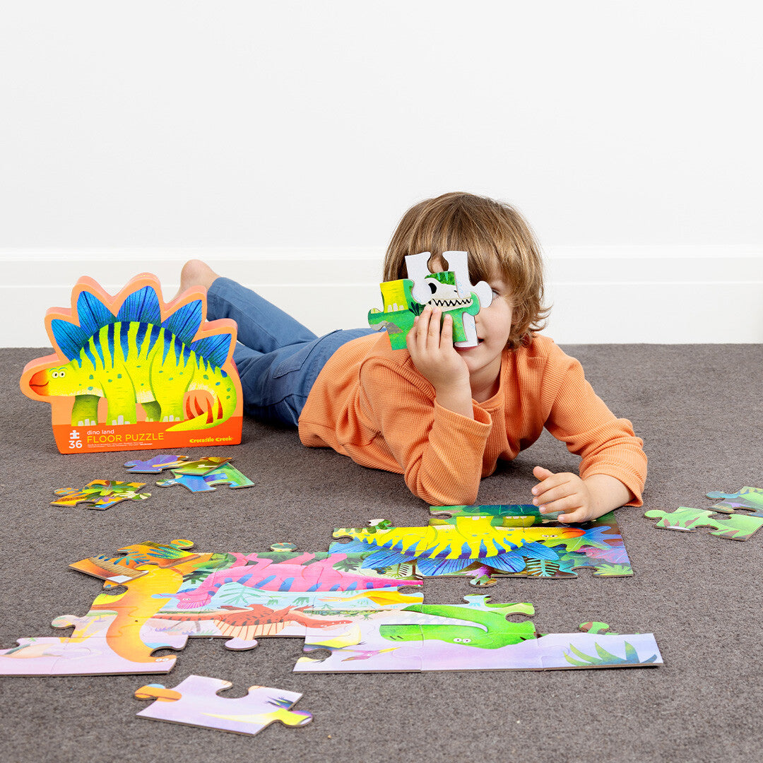 Een kind speelt met een Crocodile Creek vloerpuzzel met dinosaurussen, liggend op de grond.