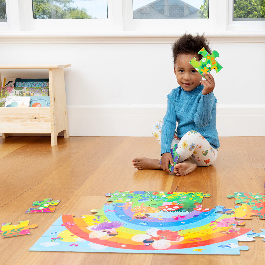 Een kind zit op de grond en houdt een puzzelstuk van de Crocodile Creek vloerpuzzel met regenboog en feeën omhoog.