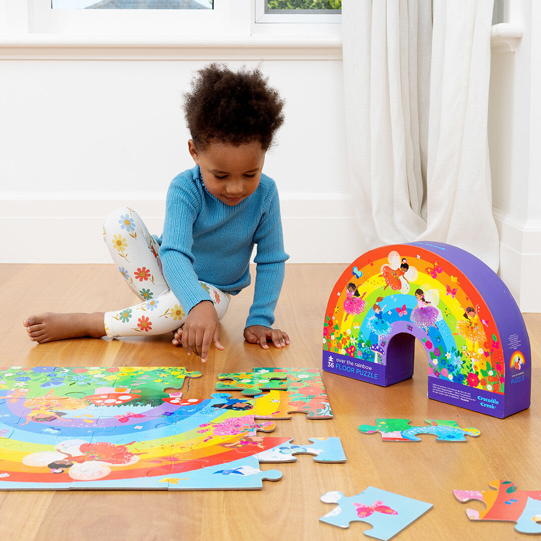 Een kind legt een puzzelstukje in de Crocodile Creek vloerpuzzel met regenboog en feeën, met de puzzeldoos ernaast.