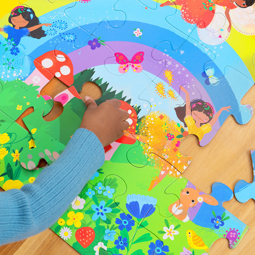 Close-up van een kinderhand die een puzzelstukje in de Crocodile Creek vloerpuzzel met regenboog en feeën plaatst.
