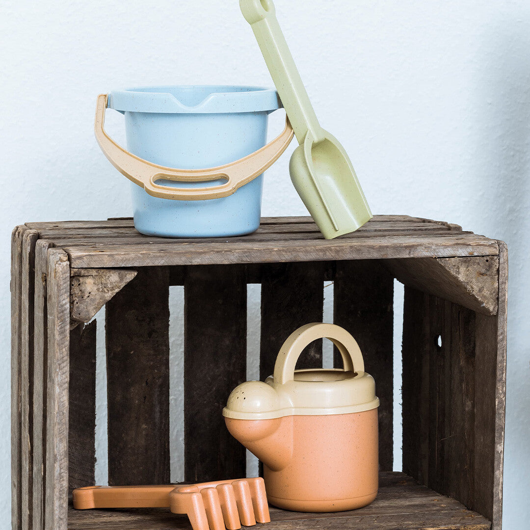 Dantoy bio-toys zandset met een emmer en schep op een houten krat, en een gieter en hark op de plank eronder.