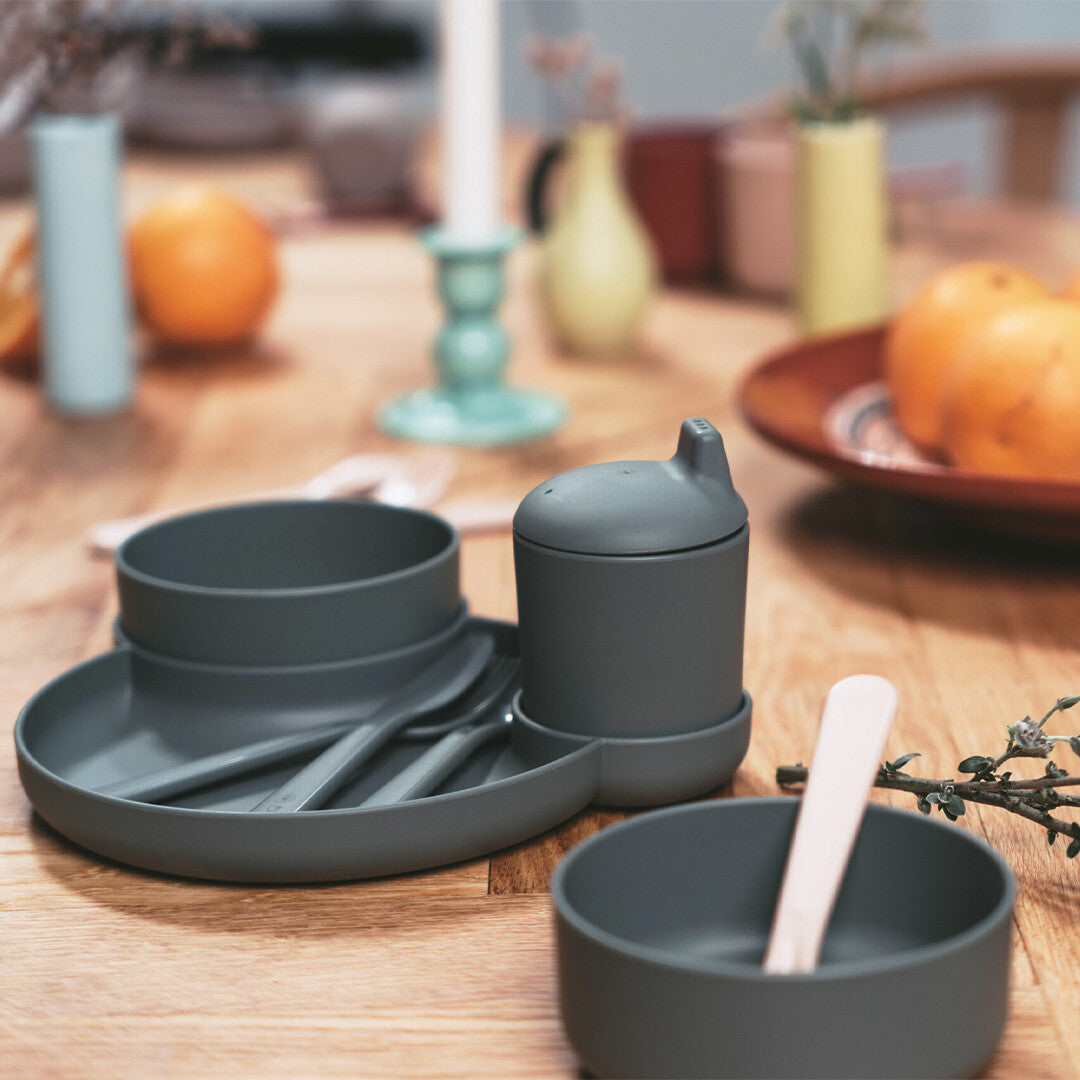 dantoy biobased kinderservies in olijfgroen op een houten tafel, met een bord, kom, beker met deksel en bestek.