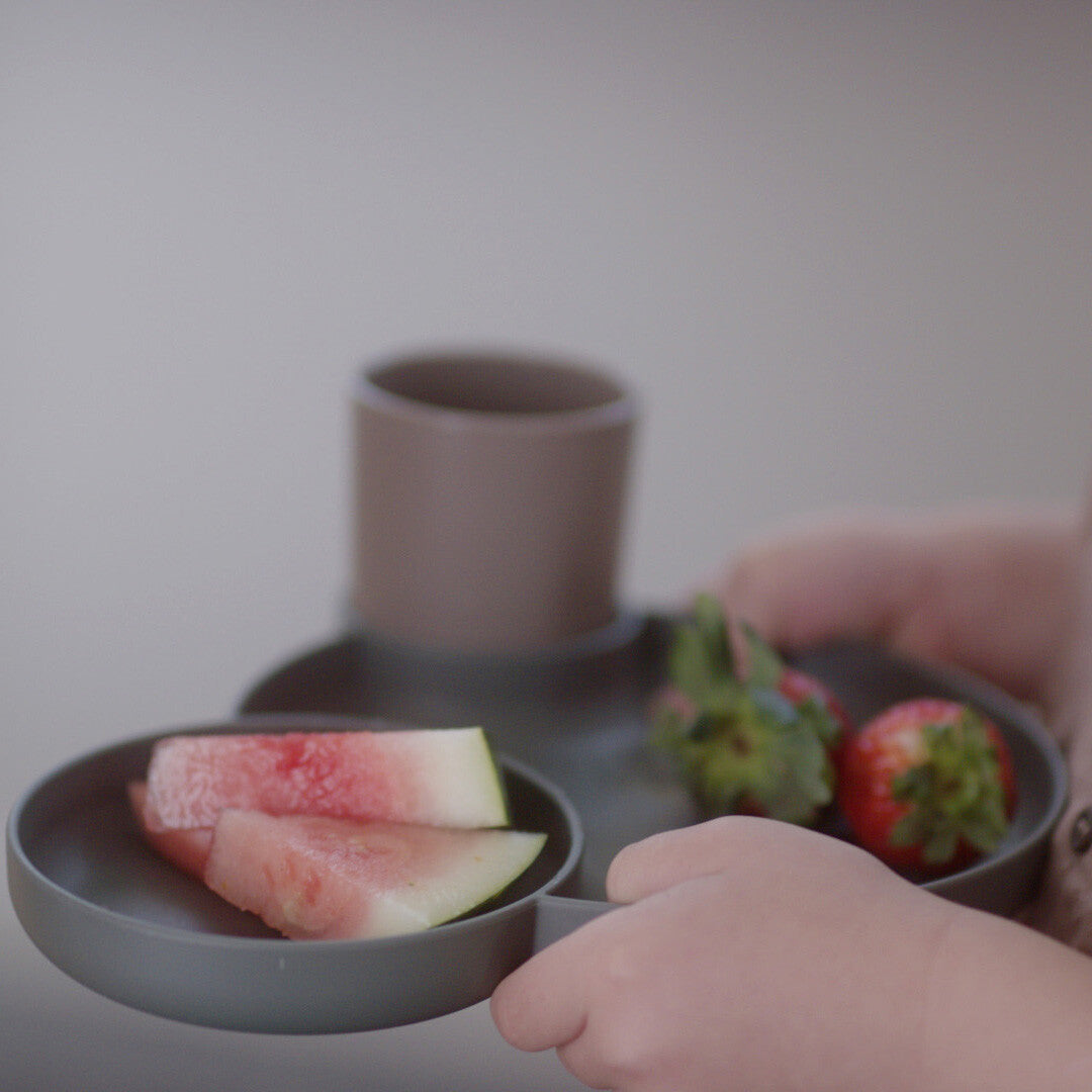 Kinderhanden houden een olijfgroen dantoy kinderserviesbord vast met watermeloen, aardbeien en drakenfruit.
