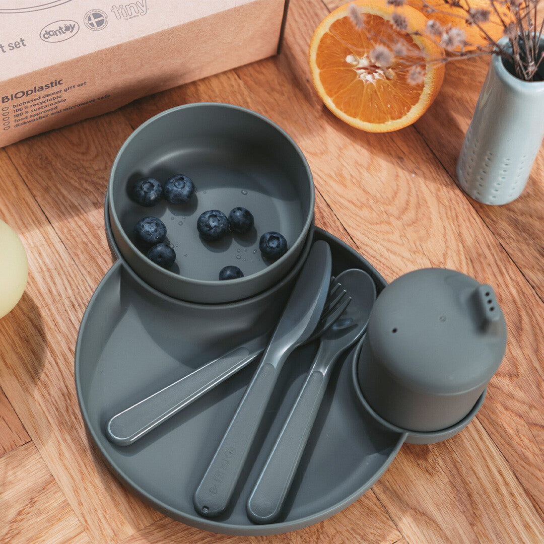 dantoy biobased kinderservies in olijfgroen op een houten tafel, met een kom gevuld met bosbessen, een bord, bestek en een beker met deksel.