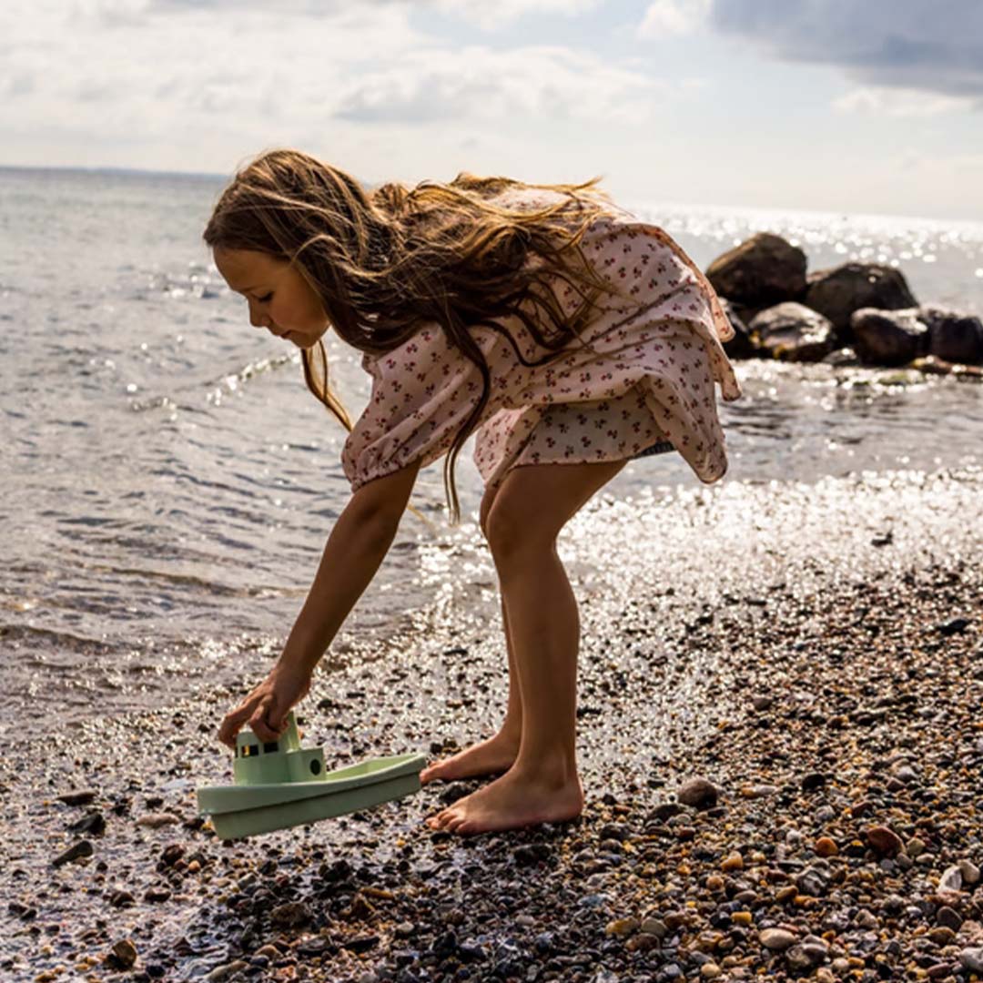 Een kind speelt met de Dantoy groene boot aan de waterkant op een kiezelstrand.