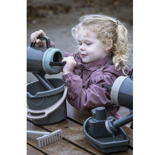 Een meisje giet water met een dantoy gieter in een emmer op een houten tafel, omringd door zandbakspeelgoed.