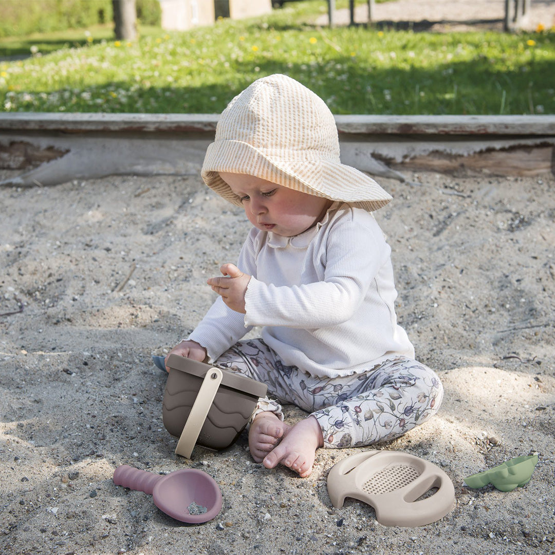 Een baby met een hoedje speelt in een zandbak met de dantoy green bean zandset.